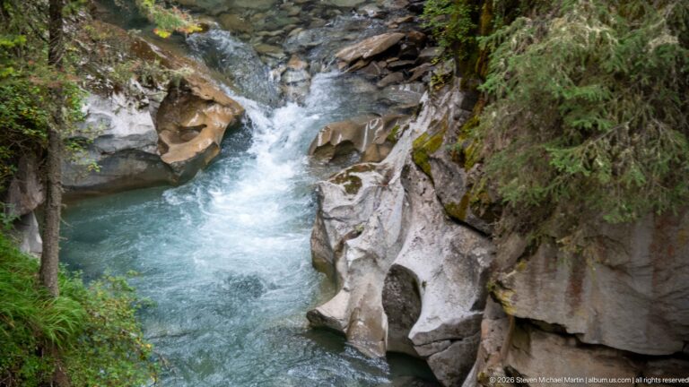 Overhead creek view shows a rounded cavity in pale rock beside blue-green flowing water.