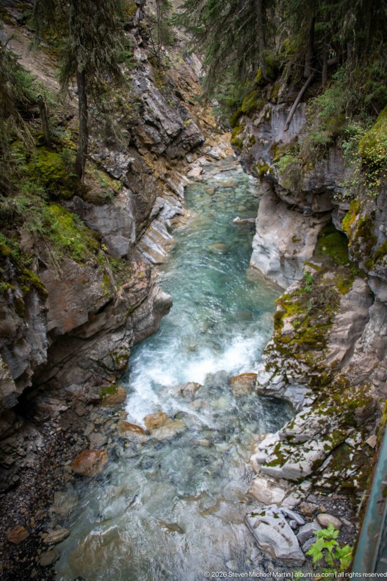 Whitewater runs through a narrow rocky channel, with a green trail railing at the lower right.