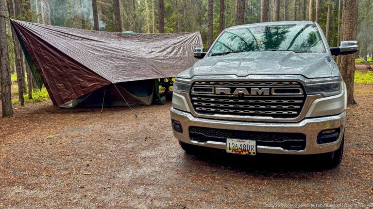 Silver RAM pickup parked in front of a tarp shelter and tent among conifers.