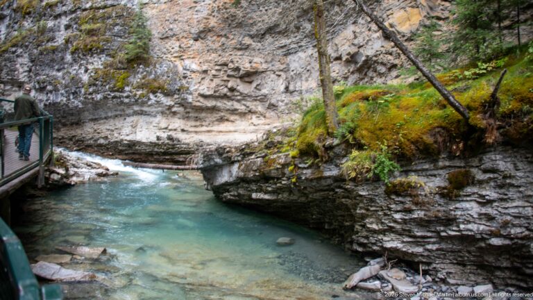 Blue-green creek pool beneath a rock shelf, with a visitor and railing at the left edge.
