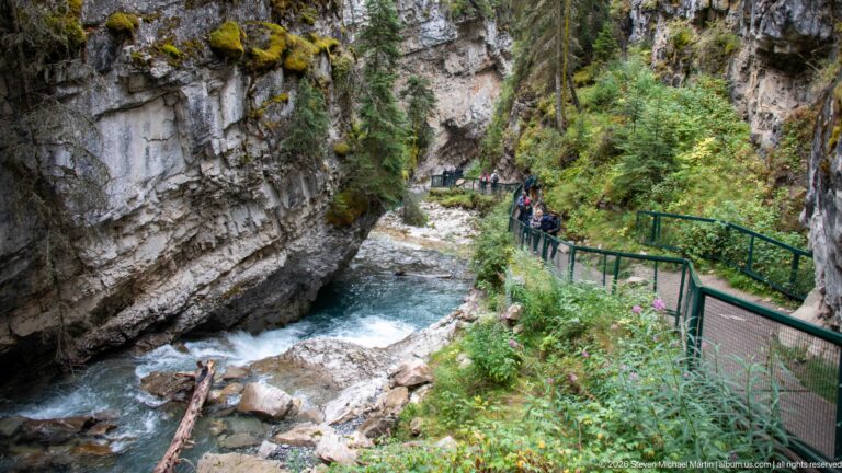 Visitors walk a green-railed path above a rocky stream in a steep forested canyon.