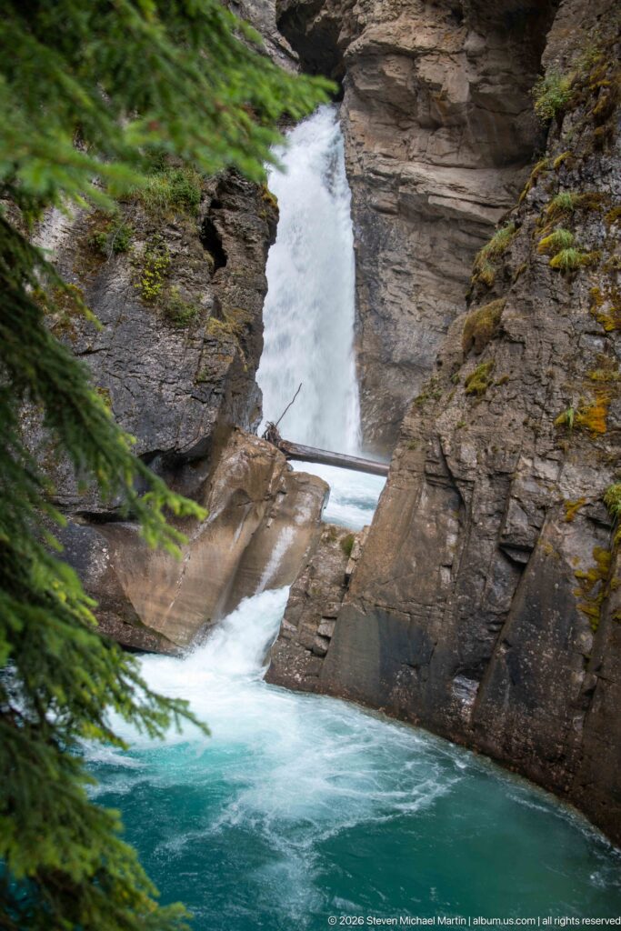 Waterfall flows beneath a wooden footbridge between steep rock walls into a blue-green pool.