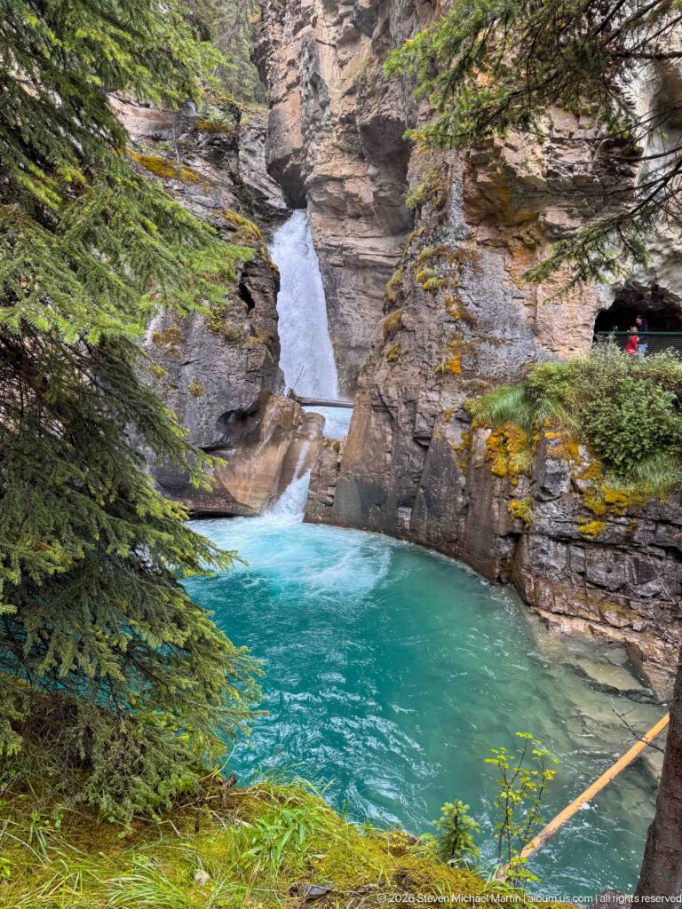 Waterfall enters a turquoise pool beside a small visitor standing in a dark rock opening.