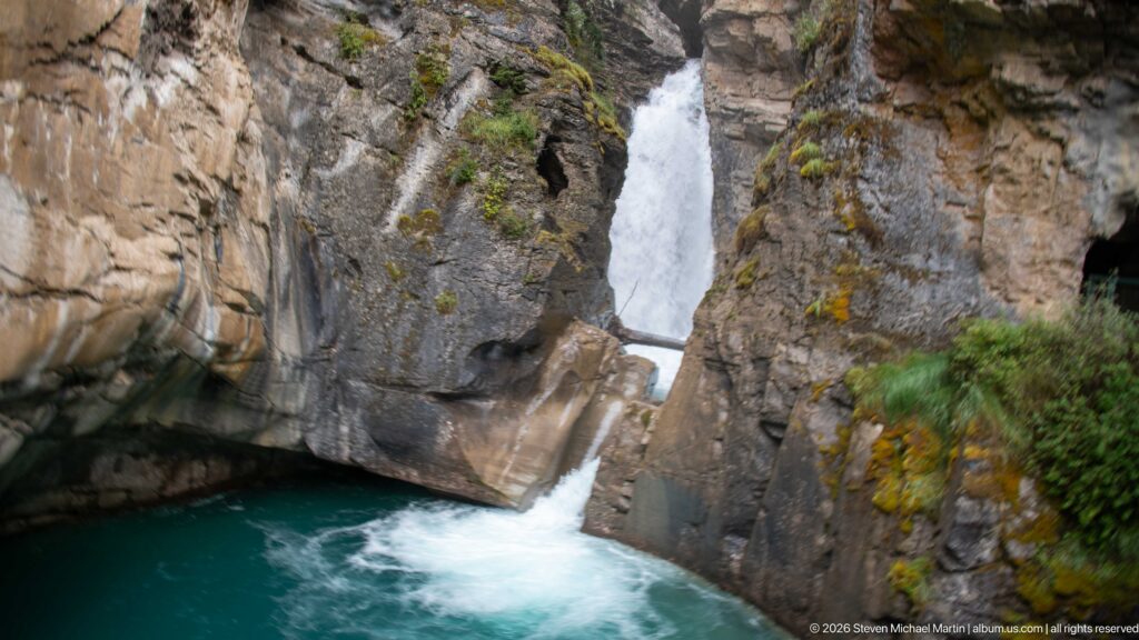 Waterfall drops through a narrow rock cleft beneath a small wooden bridge into a turquoise basin.