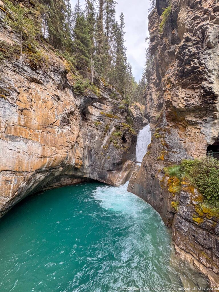Wide turquoise pool below a tall waterfall, framed by stratified rock and conifer forest.
