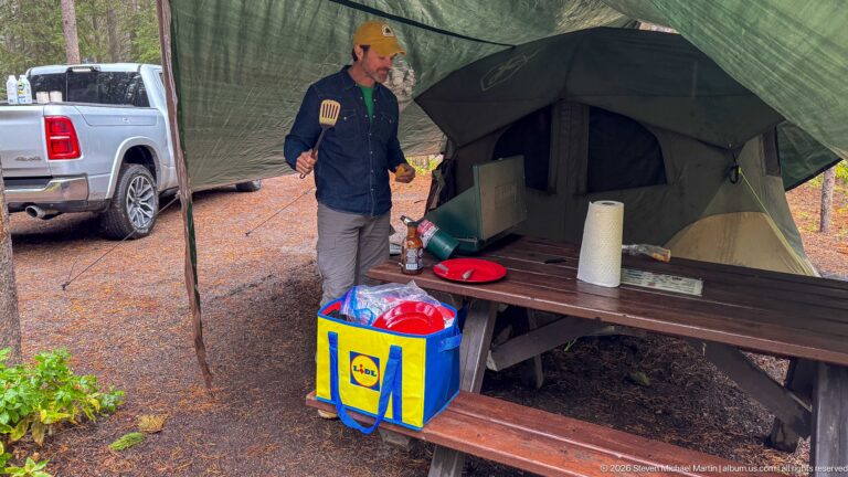 Camper with a spatula stands beside a stocked picnic table, tent, and shopping bag under a tarp.