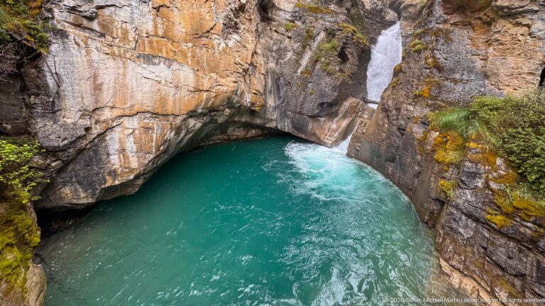 Turquoise basin beneath a sloping rock overhang, with whitewater entering from the upper right.
