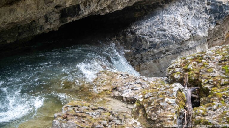 Fast creek water churns beneath a dark rock overhang and around pale stones and fallen branches.