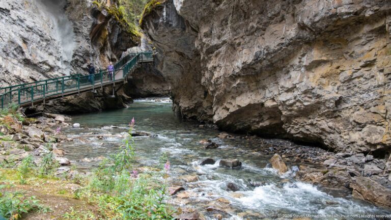 Several visitors cross a green-railed walkway above a rocky creek in a steep canyon.