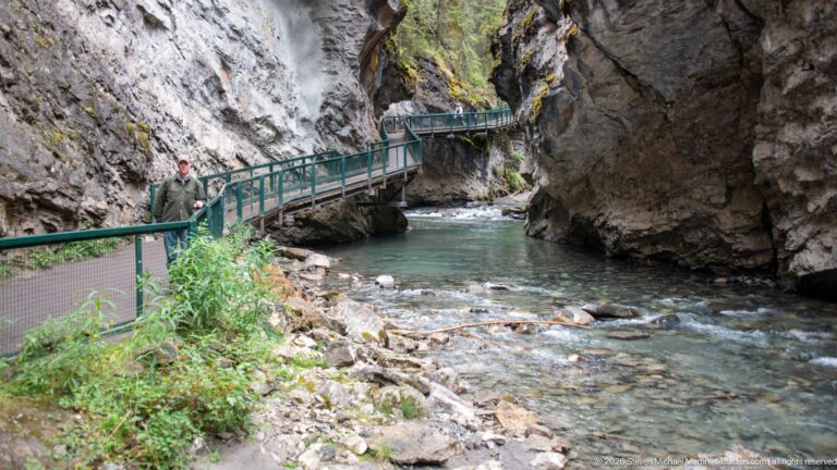Visitor in a green jacket stands on the walkway above the creek, framed by rock walls and railings.
