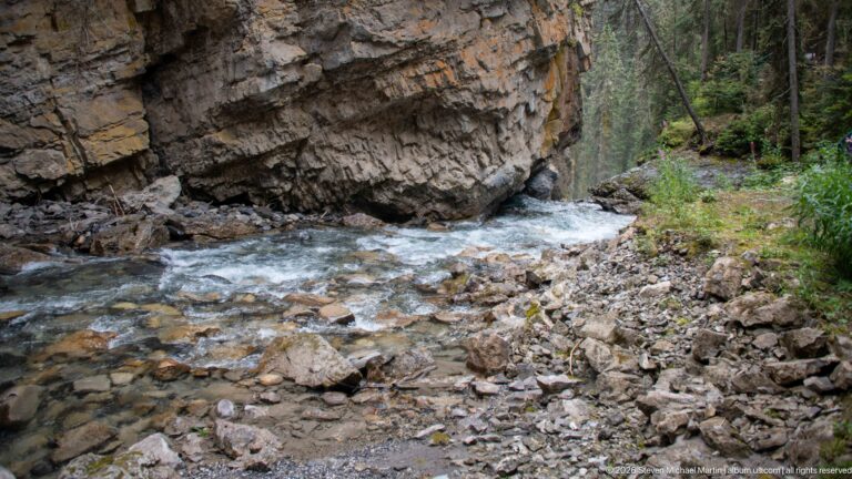Shallow creek passes beneath a projecting rock ledge beside a gravel and cobble bank.