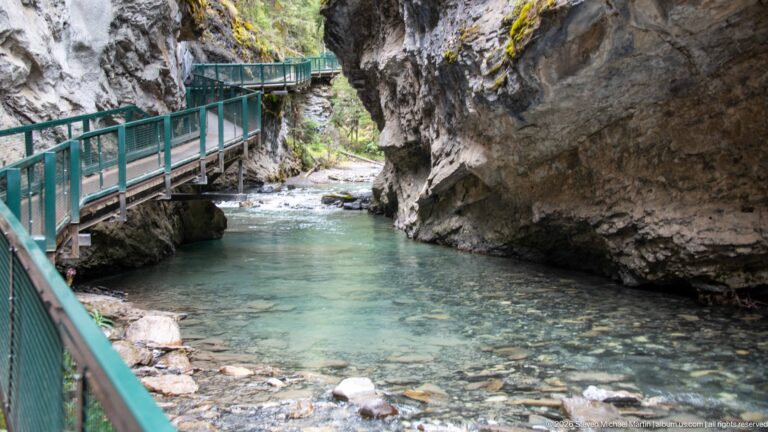 Green walkway curves beside a turquoise pool beneath a projecting rock overhang.