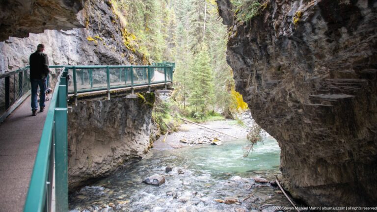 A lone hiker crosses a green-railed walkway above the creek in a narrow canyon.