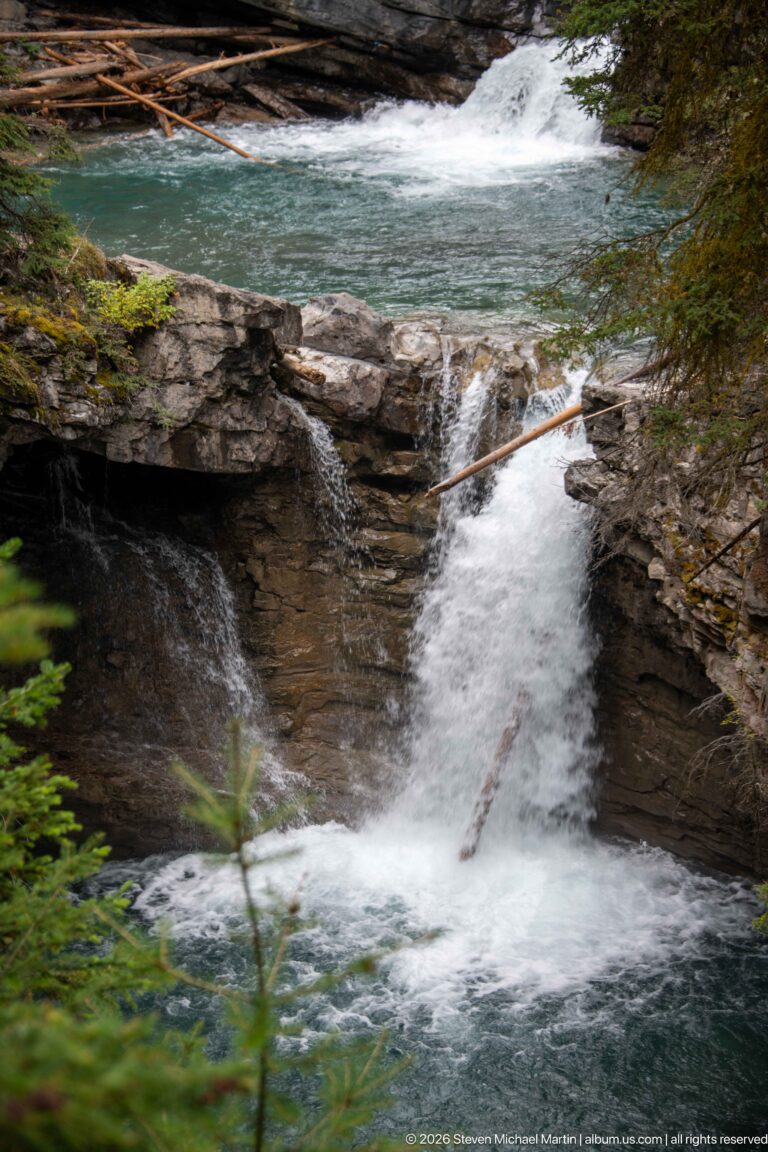 Blue-green pool spills over a low rock ledge into a short cascade, with a branch across the foreground.