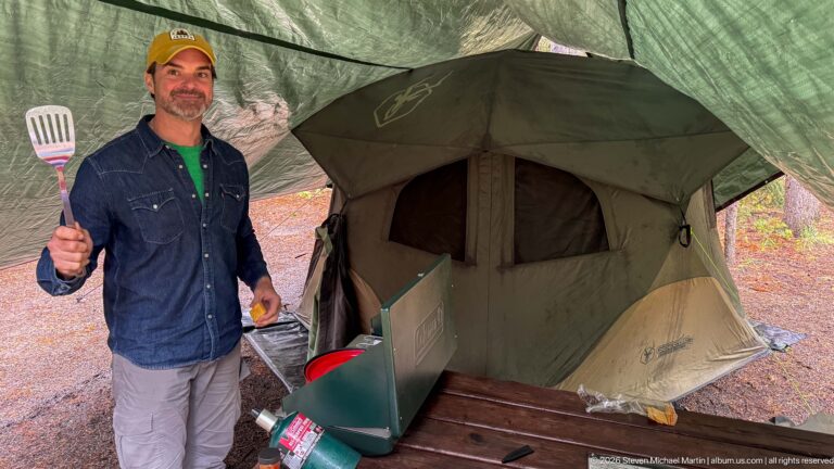 Camper holds a spatula beside an open tent and a table set with cooking supplies.