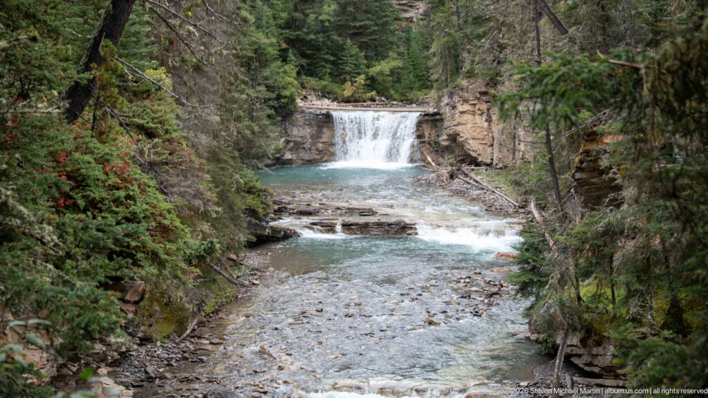 Shallow creek leads through a wooded rock corridor toward a waterfall in the distance.