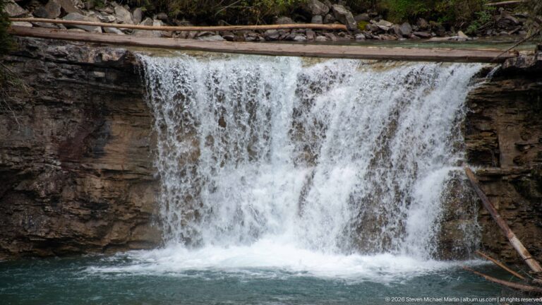 Wide sheet of water pours over a level rock ledge into a shadowed pool.