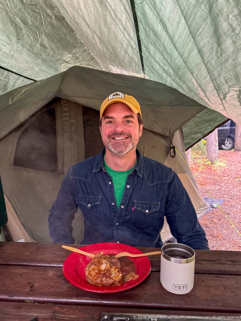 Camper in an orange cap sits at a picnic table with a meal beneath a green tent rainfly.