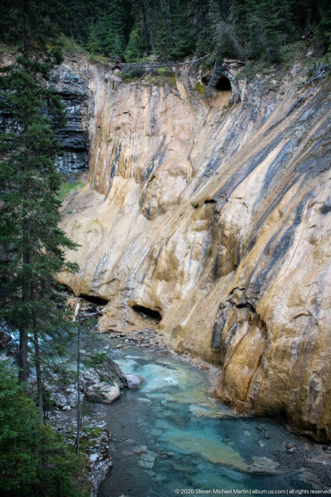 Blue-green creek beneath a pale sloping rock face with dark horizontal layers at the edge.