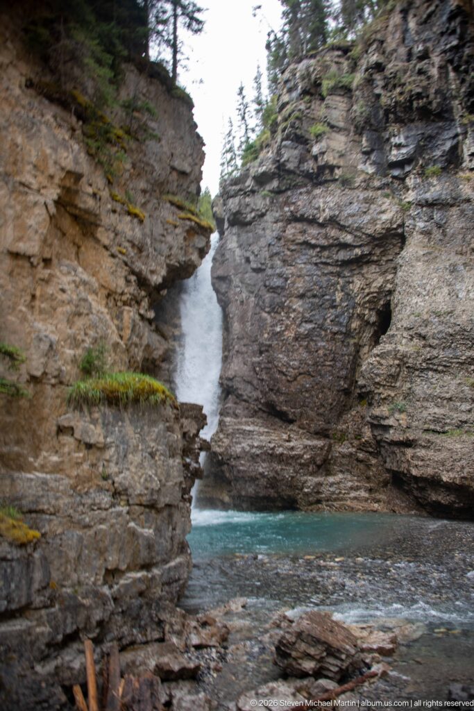 A waterfall and turquoise pool seen through a narrow cleft between tall canyon walls.
