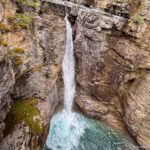 A slender waterfall descends between warm-toned rock ledges into a turquoise pool.