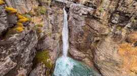 A slender waterfall descends between warm-toned rock ledges into a turquoise pool.