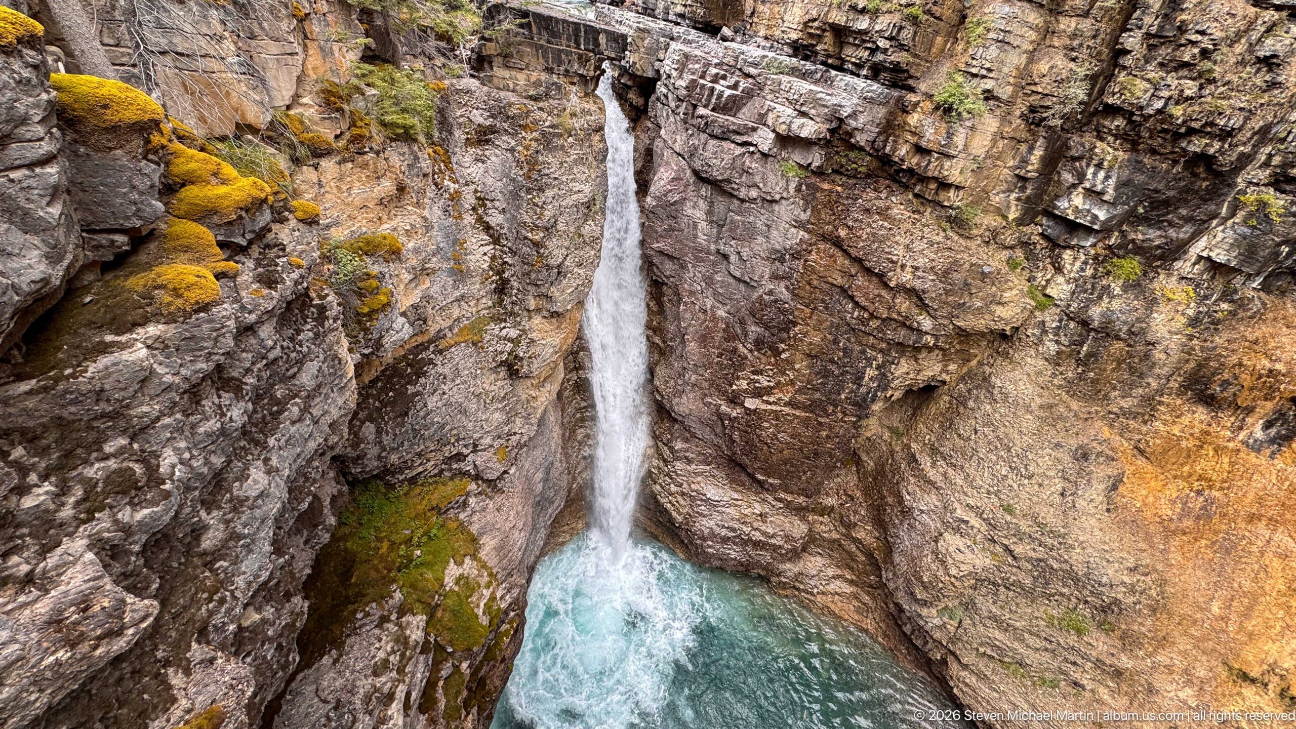 A slender waterfall descends between warm-toned rock ledges into a turquoise pool.