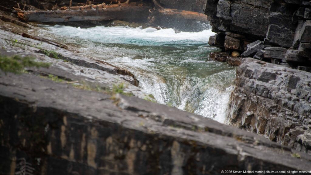 Turbulent blue-green creek water spills over a ledge beyond a dark stone parapet.