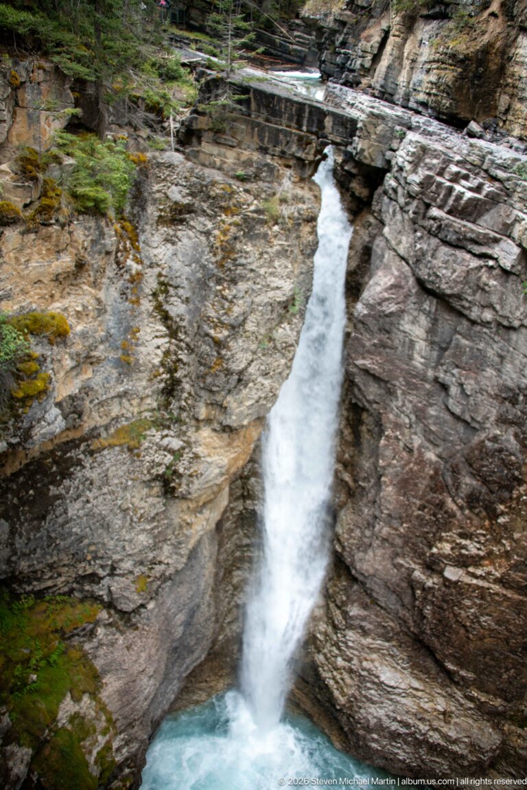 A narrow waterfall plunges between layered rock walls into a turquoise basin.