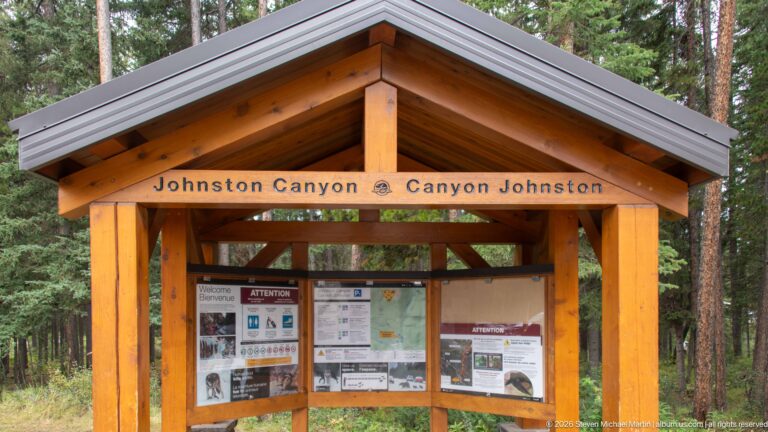 Wooden Johnston Canyon information kiosk with a peaked roof and posted trail notices, surrounded by conifers.