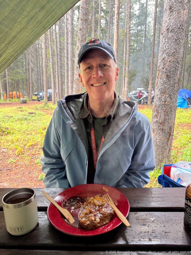 Smiling camper sits beneath a green tarp with a plate of food and metal mug on the table.