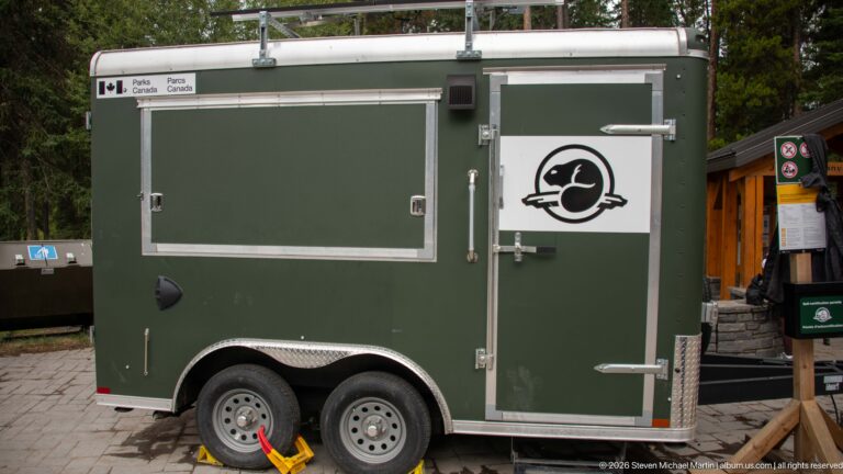 Green Parks Canada trailer with a white beaver-emblem panel, parked beside a wooden building.