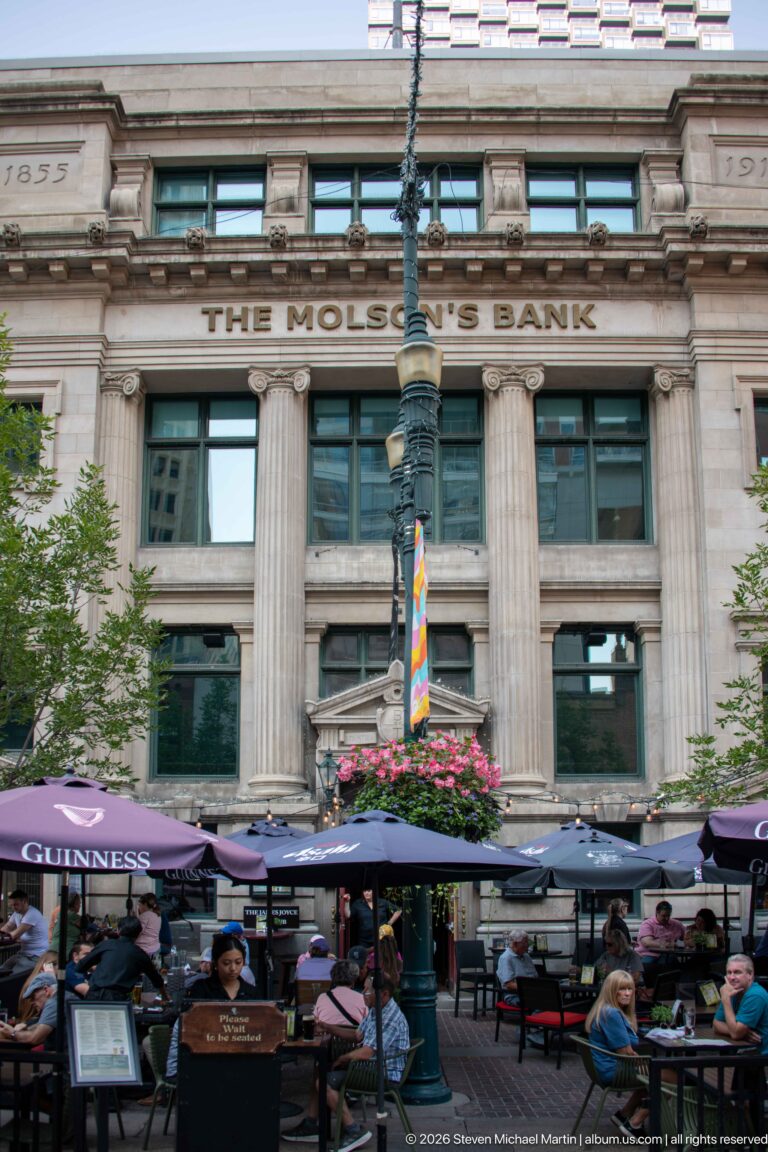Former Molson’s Bank classical stone façade behind restaurant patios and hanging flowers in downtown Calgary.