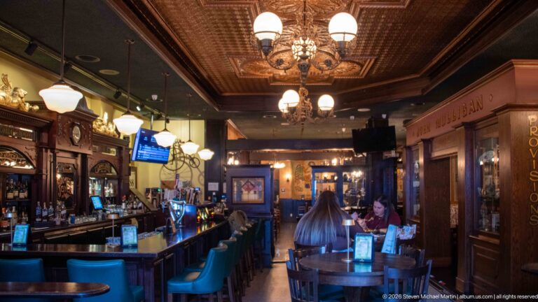 Long James Joyce pub interior with teal bar stools, chandeliers, patterned ceiling, carved woodwork, and dining tables.