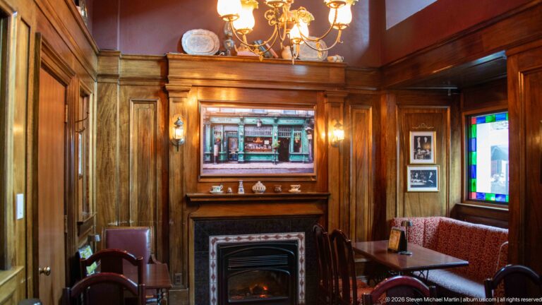 Wood-paneled Irish pub room with fireplace, red upholstered seating, framed pictures, and stained-glass window.