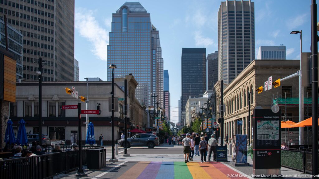 Downtown Calgary street with rainbow pavement, pedestrians, restaurant patios, historic buildings, and high-rise towers.
