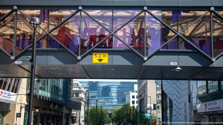 Glass-enclosed pedestrian bridge with diagonal framing crossing above a downtown Calgary street and 5.0-metre clearance sign.