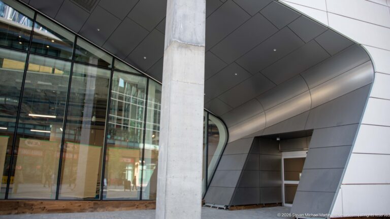 Curved silver metal entrance wall beside glass windows and a large concrete column on a modern Calgary building.