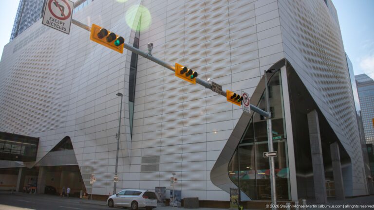 Large pale faceted building with textured metal cladding and a tall angular glass entrance at a Calgary intersection.