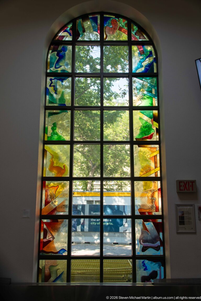 Large arched window with colorful abstract stained-glass border surrounding clear central panes and a view of trees.