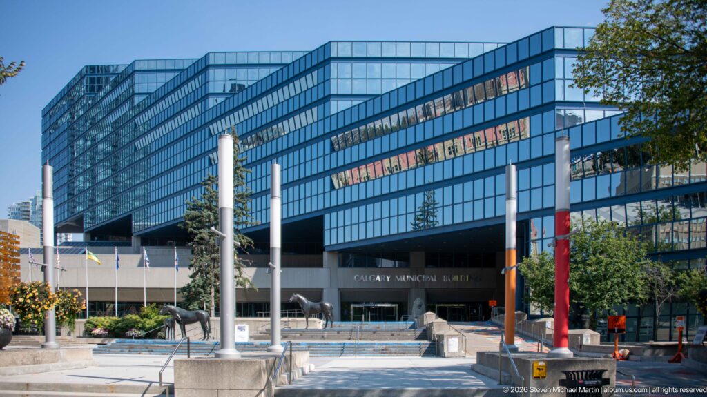 Calgary Municipal Building entrance beneath a blue-glass office block, with columns, flags, sculptures, and plaza landscaping.