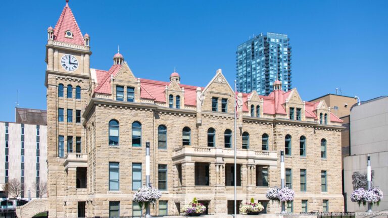Historic Calgary City Hall with sandstone façade, red roofs, clock tower, flowers, and a modern high-rise behind.