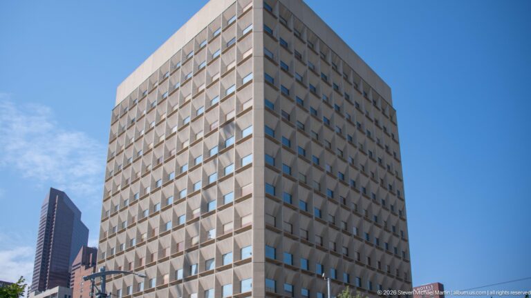 Pale rectangular Calgary high-rise with a strict grid of recessed square windows under a blue sky.