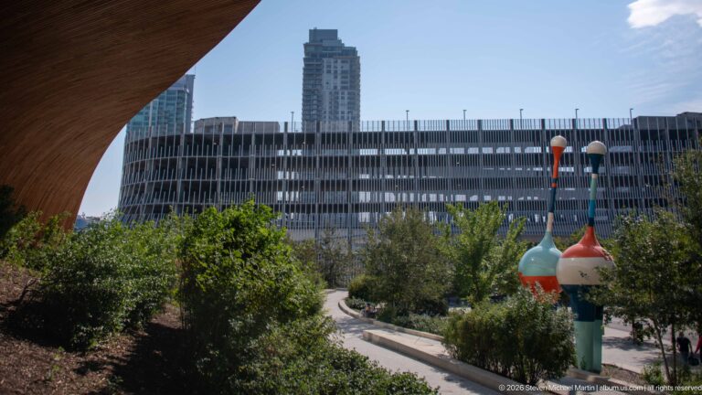 Landscaped path, shrubs, colorful pole-mounted forms, and modern buildings beside Calgary Central Library.