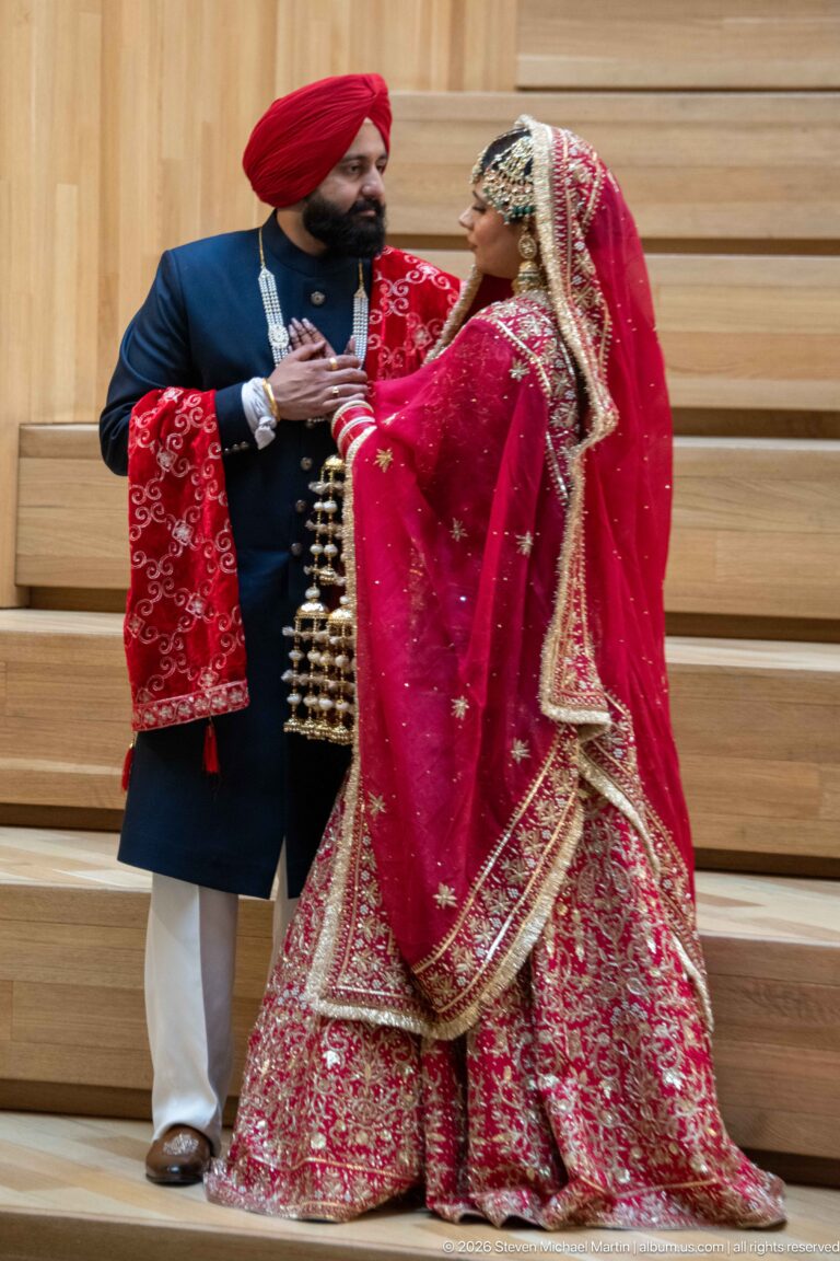 South Asian wedding couple in elaborate red, gold, and dark formal clothing posing on wide wooden stairs.