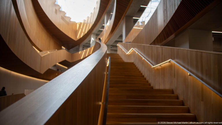 Wide wooden staircase rising beneath curving wood-clad terraces inside Calgary Central Library.