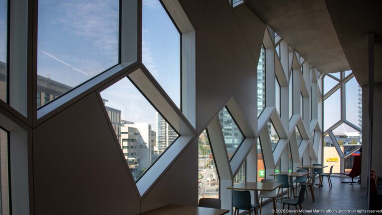 Tables and chairs beside repeating angular windows inside Calgary Central Library, with city buildings visible outside.