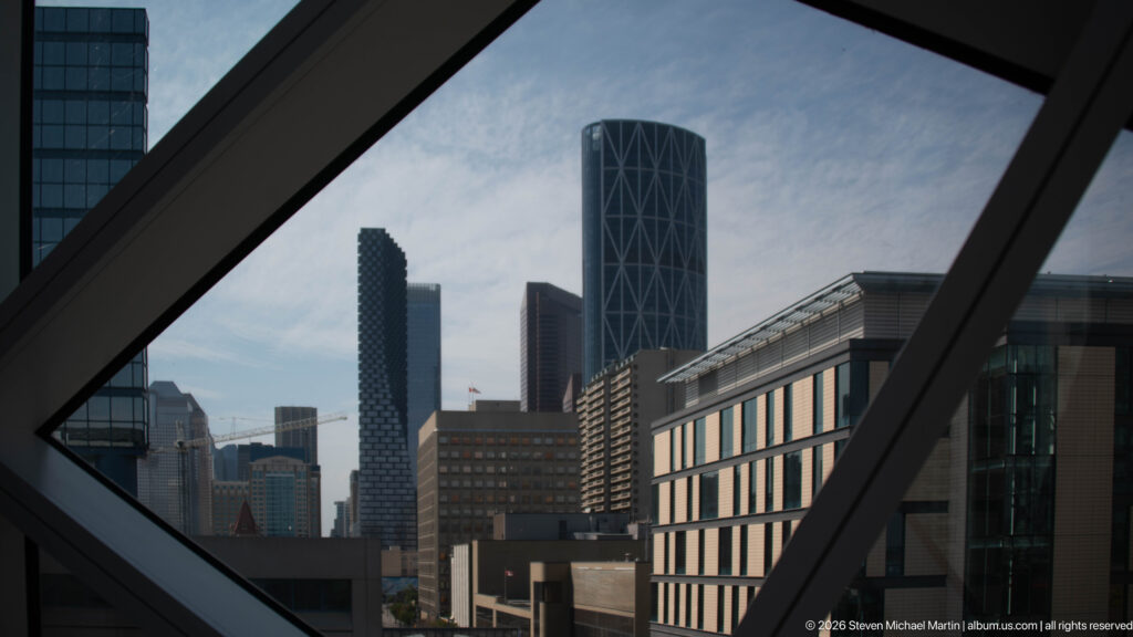 Downtown Calgary towers seen through dark diagonal window framing inside Calgary Central Library.