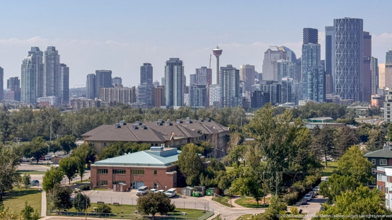 Downtown Calgary skyline with Calgary Tower and high-rises beyond trees, low buildings, and neighborhood streets.