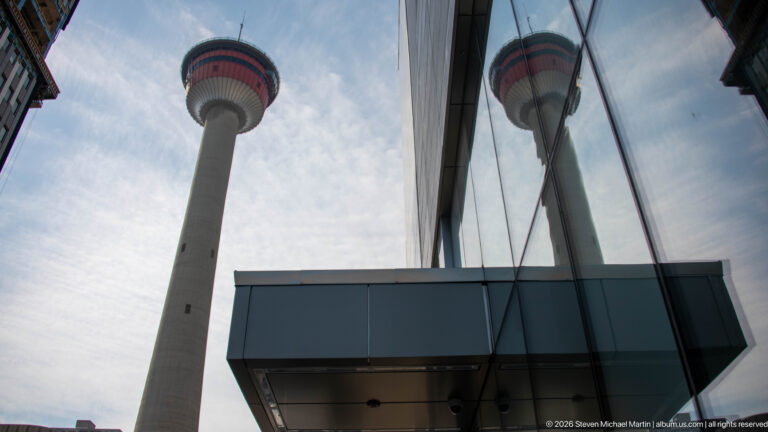 Calgary Tower and its reflection in a glass building, viewed upward beside a dark projecting canopy.